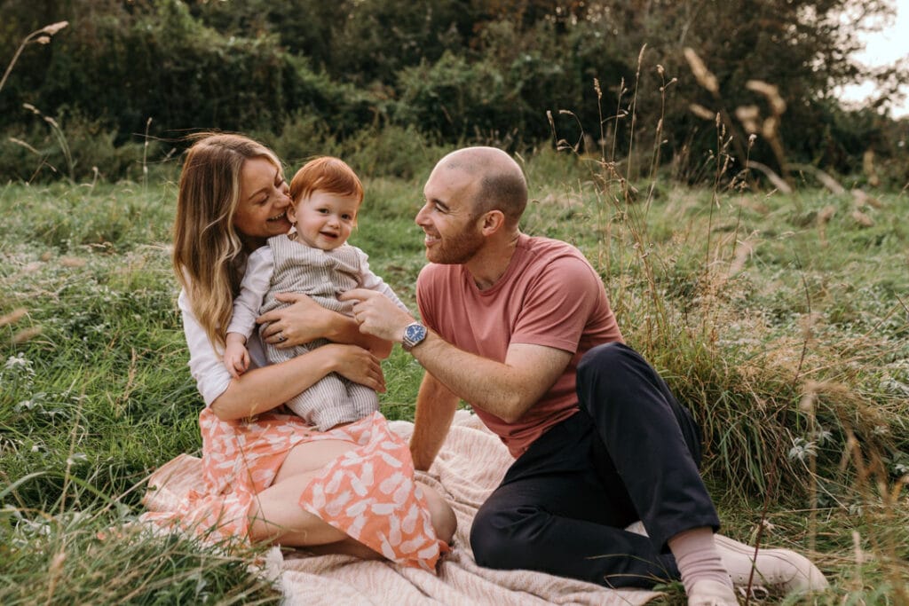 mum is cuddling her boy and looking at him and dad is looking at his son too. They are sitting on the blanket in the field. Summer family photo session in Hook, Hampshire. Ewa Jones Photography