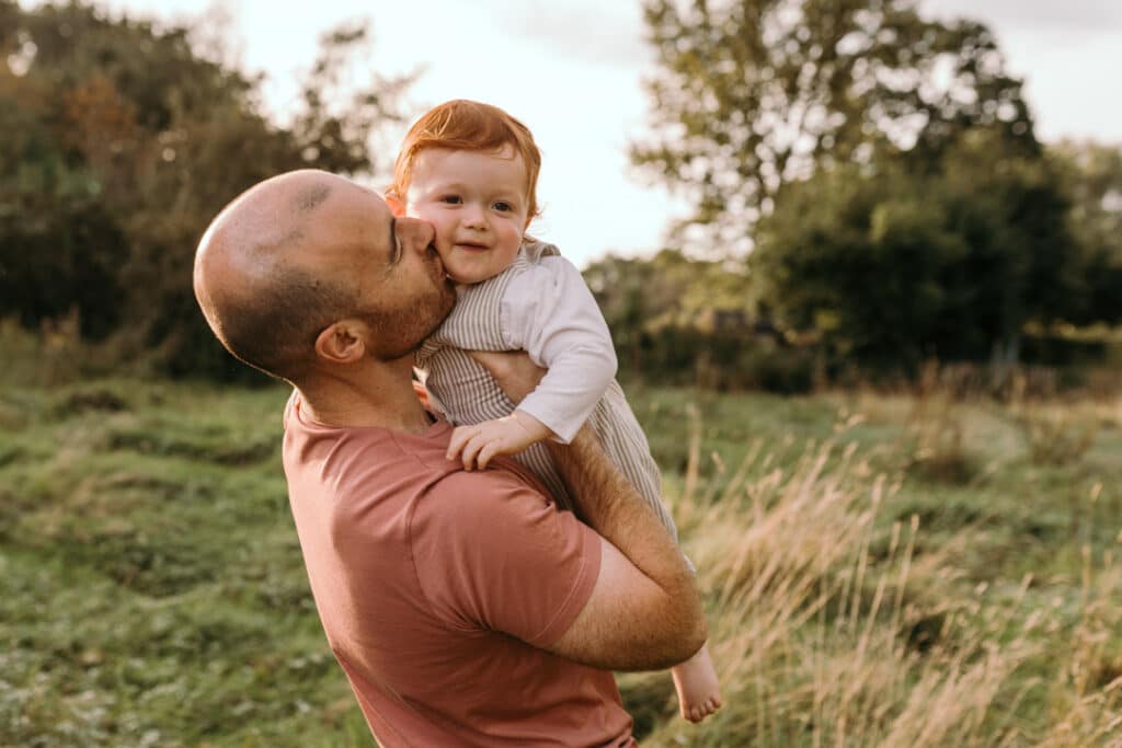 dad is kissing his baby boy. Dad is wearing pink top and boy is wearing stripy outfit. They are in the field of long grass. family photographer in Hook, Hampshire. Ewa Jones Photography