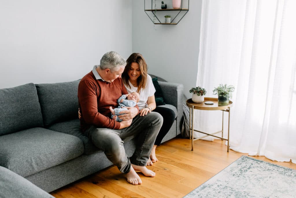 Mum and dad are sitting on the sofa and looking down at their newborn baby boy. In home natural lifestyle newborn photo session. Ewa Jones Photography