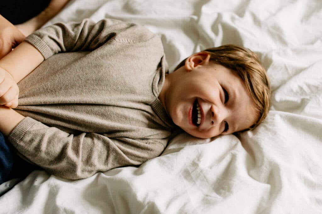 Little boy is laying on bed and being tickled by his dad. the boy is laughing. family photo shoot in Basingstoke, Hampshire. Ewa Jones Photography