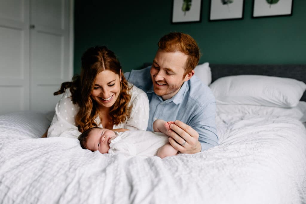 Mum and dad are laying on the bed and looking down on their newborn baby girl. She is wearing baby grow and mum is wearing lovely dress. Newborn photography in Hampshire. Ewa Jones Photography