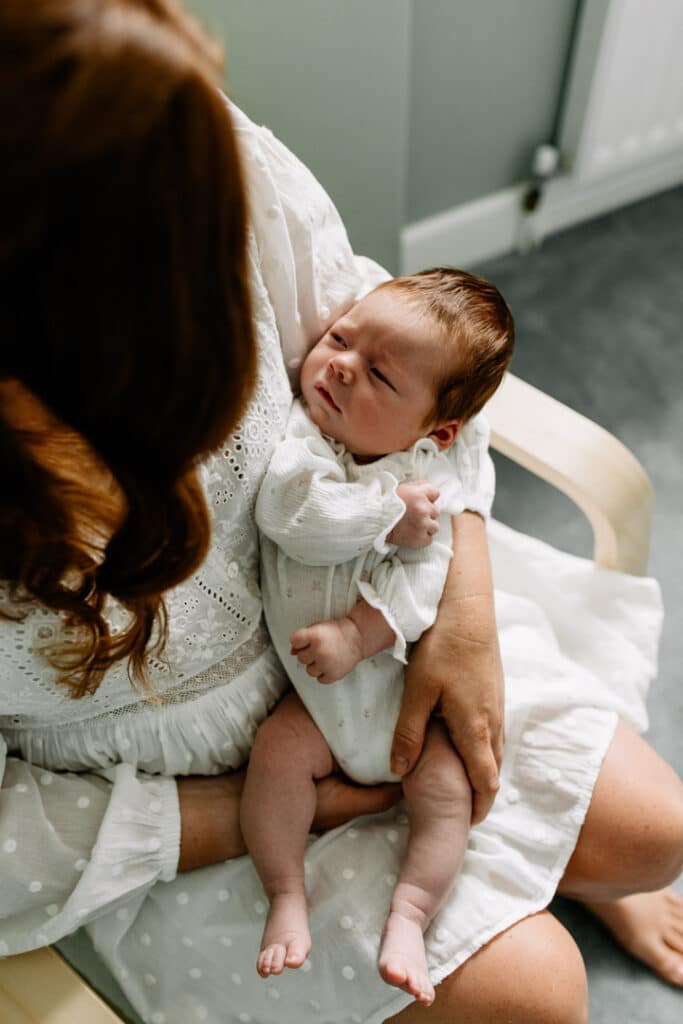 Mum is sitting in newborn baby nursery and holding her little girl. Newborn girl is looking up at her mum. Natural newborn photographer in Basingstoke, Hampshire. Ewa Jones Photography