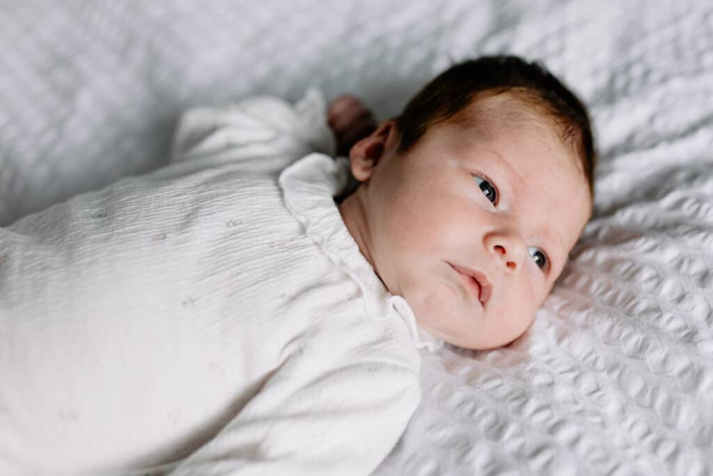 Newborn baby girl is laying on bed and looking away. she is wearing lovely white baby grow. Newborn photographer in Basingstoke, Hampshire. Ewa Jones Photography