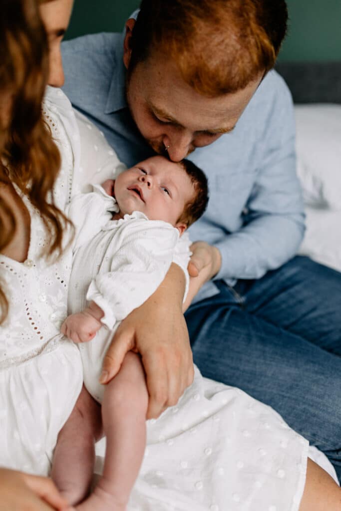 Mum and dad are sitting on the edge of the bed and mum is holding her newborn baby girl. Dad is kissing baby on her head. Lifestyle newborn photo shoot in Hampshire. Ewa Jones Photography