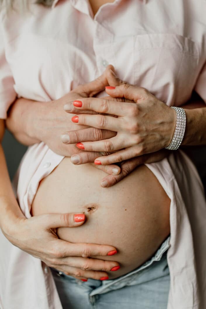 Pregnant mum is wearing pink shirt and holding her baby bump. Dad is standing behind mum and has his hands wrapped around mum waist. Lifestyle maternity photo shoot in Hampshire. Ewa Jones Photography