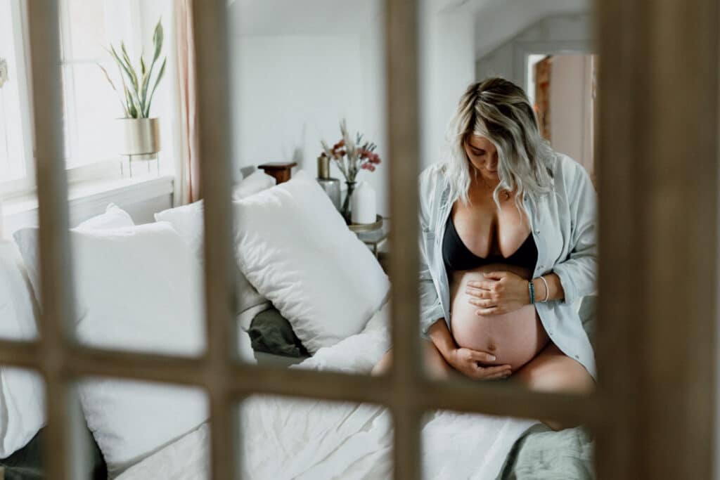 Expecting mum is holding her bump and sitting on the bed. She is wearing a blue jean shirt and a black bra. What to wear for a maternity photo session. Maternity photographer in Hampshire. Ewa Jones Photography