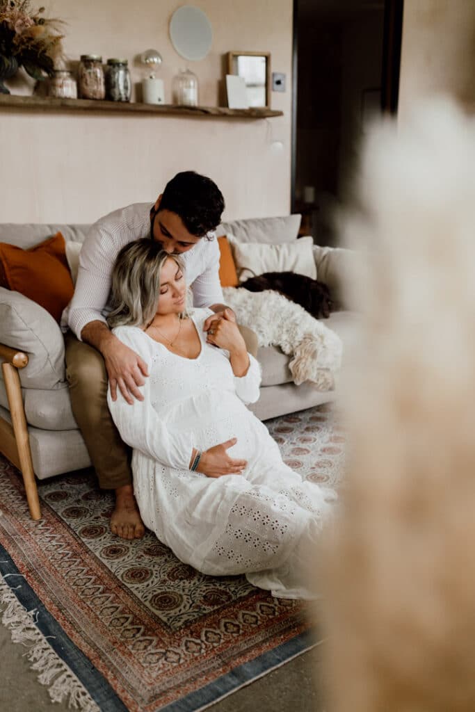 Expecting parents are relaxing in the living room. Dad is sitting on the sofa and mum is sitting in front of him on the floor. Lovely maternity photo shoot in Hampshire. Ewa Jones Photography