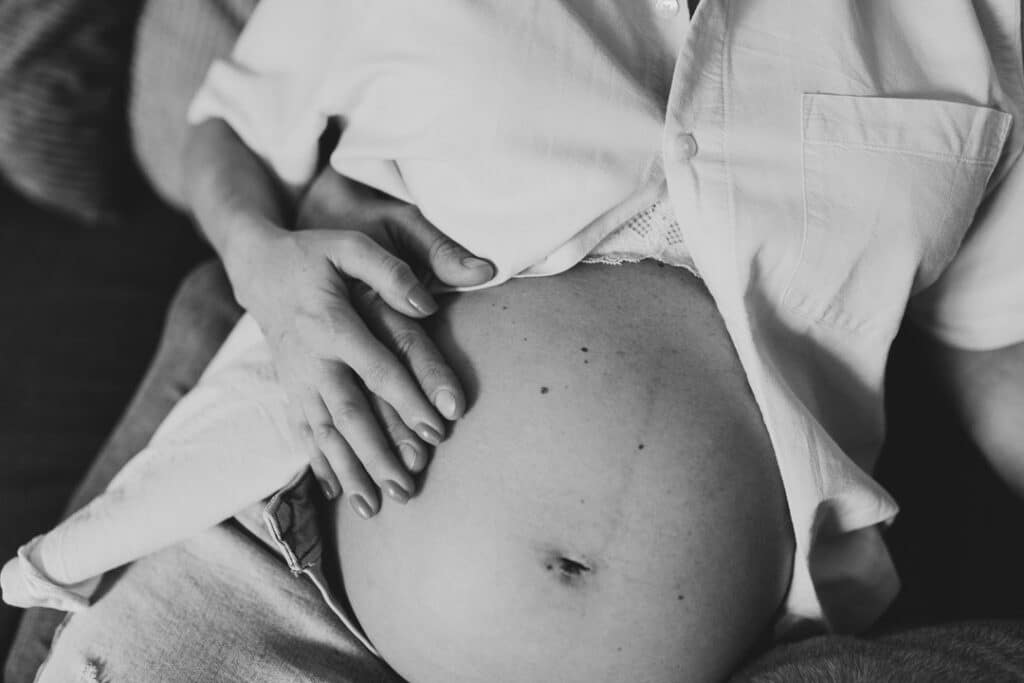Black and white image of baby bump. Mum is sitting on the sofa and has her hand on her bump. Dad is also holding baby bump. Maternity photographer in Basingstoke, Hampshire. Ewa Jones Photography