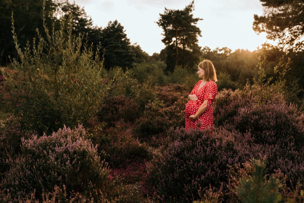 Expecting mum is standing in the woods and holding her baby bump. She is wearing lovely red dress with white flowers. Maternity photographer in Basingstoke, Hampshire. Ewa Jones Photography