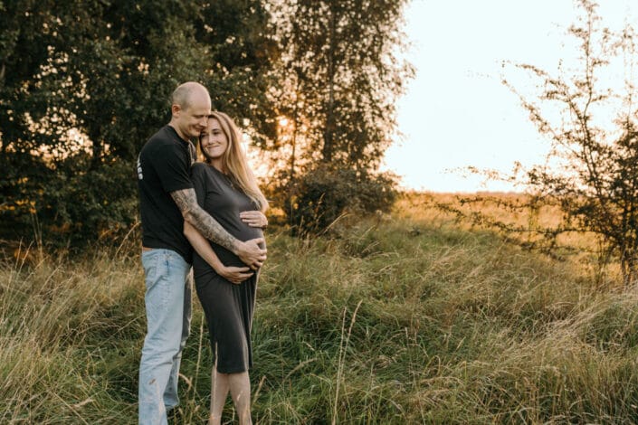 Expecting parents are standing in the fields and cuddling up to each other. Mum and dad are holding bump. Maternity photographer in Basingstoke, Hampshire. Ewa Jones Photography