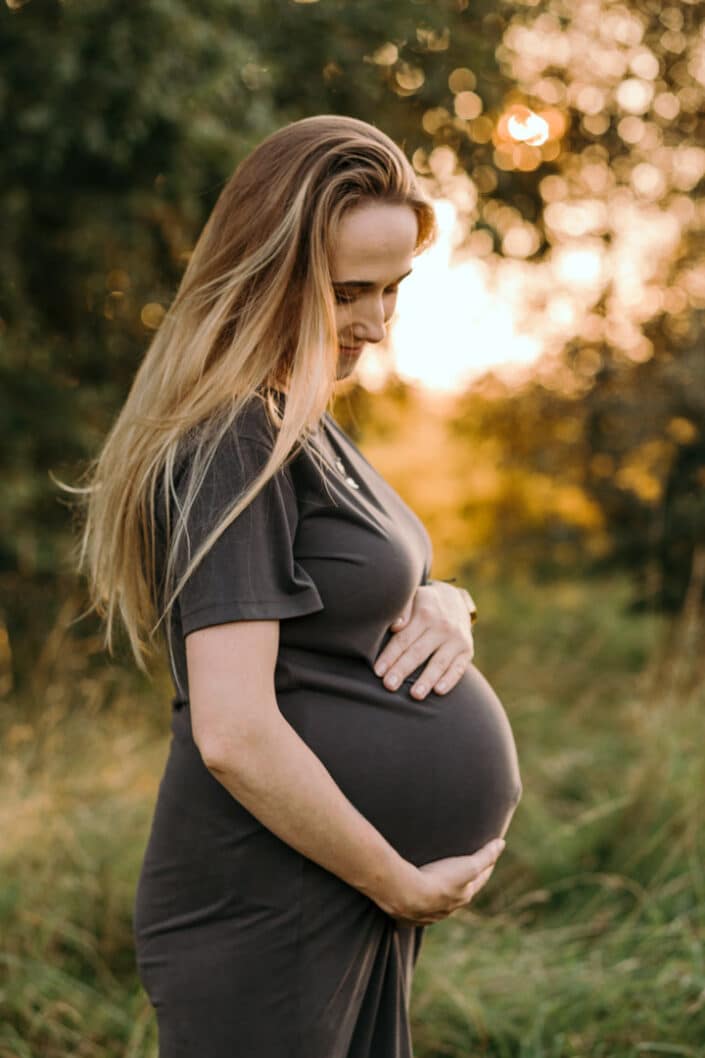 Pregnant mum is looking down at her baby bump and holding her baby bump. She is wearing lovely brown dress and the photo session is during golden hour. Maternity photographer in Hampshire. Ewa Jones Photography