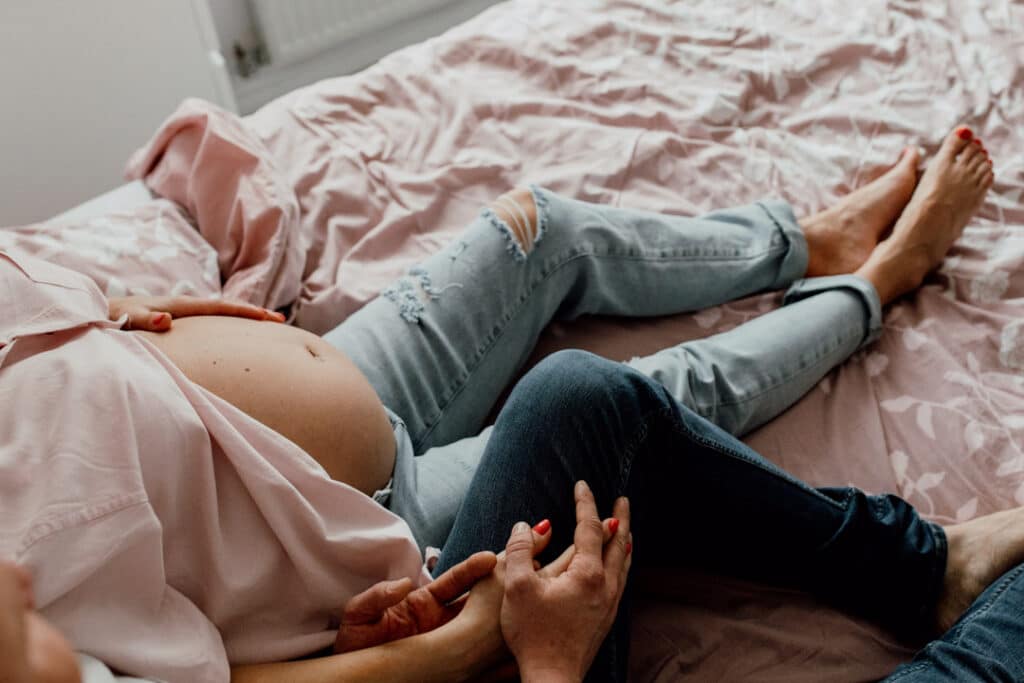 Pregnant mum is sitting on the bed with her husband on her side. She is holding baby bump and mum and dad are interlocking their fingers. Lovely natural lifestyle maternity photo shoot in Hampshire. Ewa Jones Photography