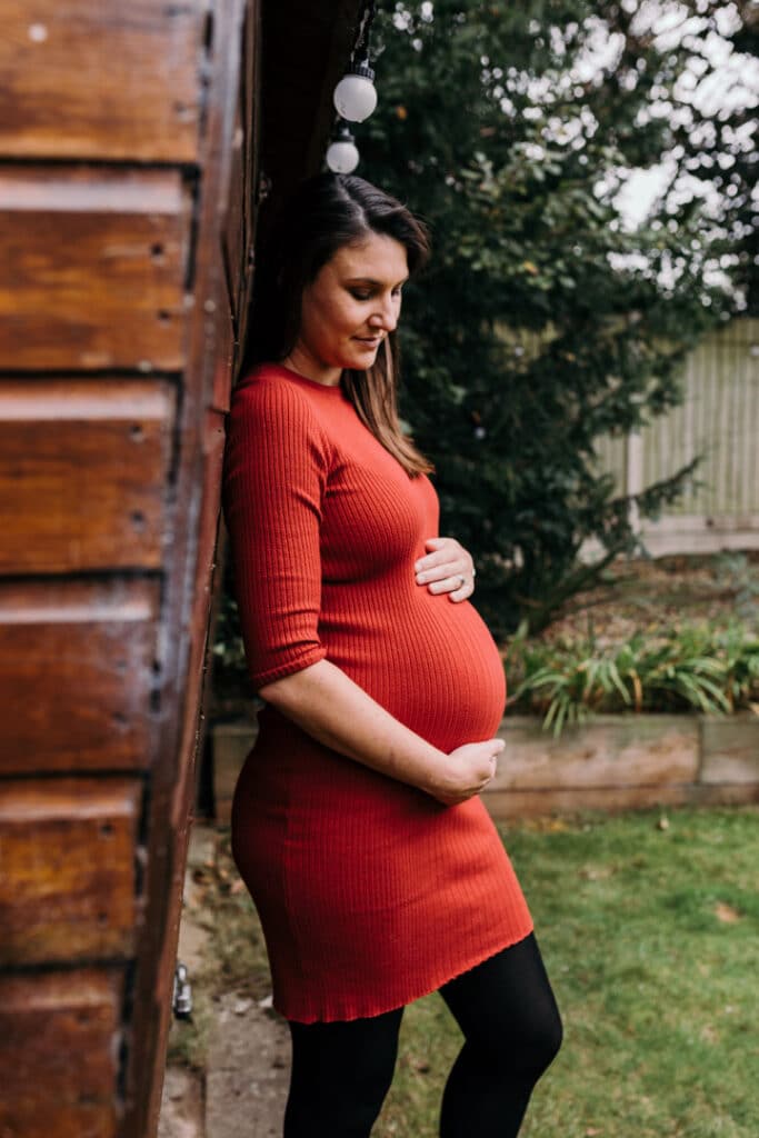 Pregnant mum is wearing lovely red dress and holding her baby bump. She is standing in the garden. Maternity photo shoot in London. Ewa Jones Photography