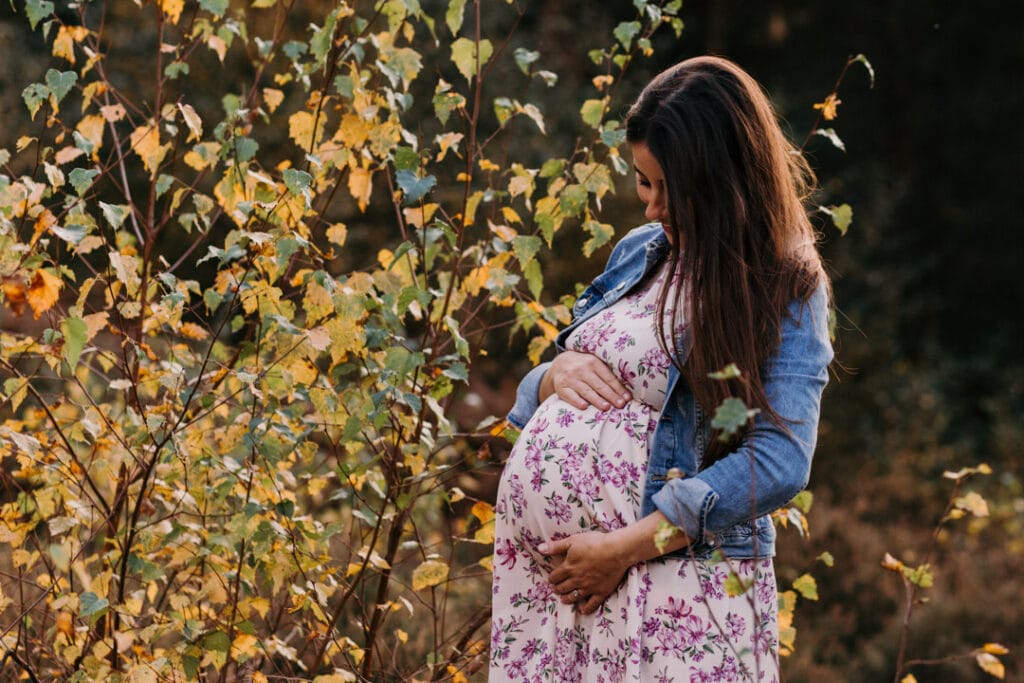 Expecting mum is holding her baby bump. She is wearing lovely flower dress and a jean jacket. Maternity photographer in Hampshire. Ewa Jones Photography