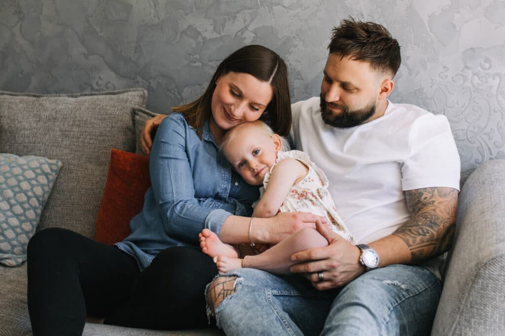 Expecting mum and dad are sitting on the sofa. their older daughter is cuddling up to her mum. Maternity photographer in Basingstoke. Ewa Jones Photography