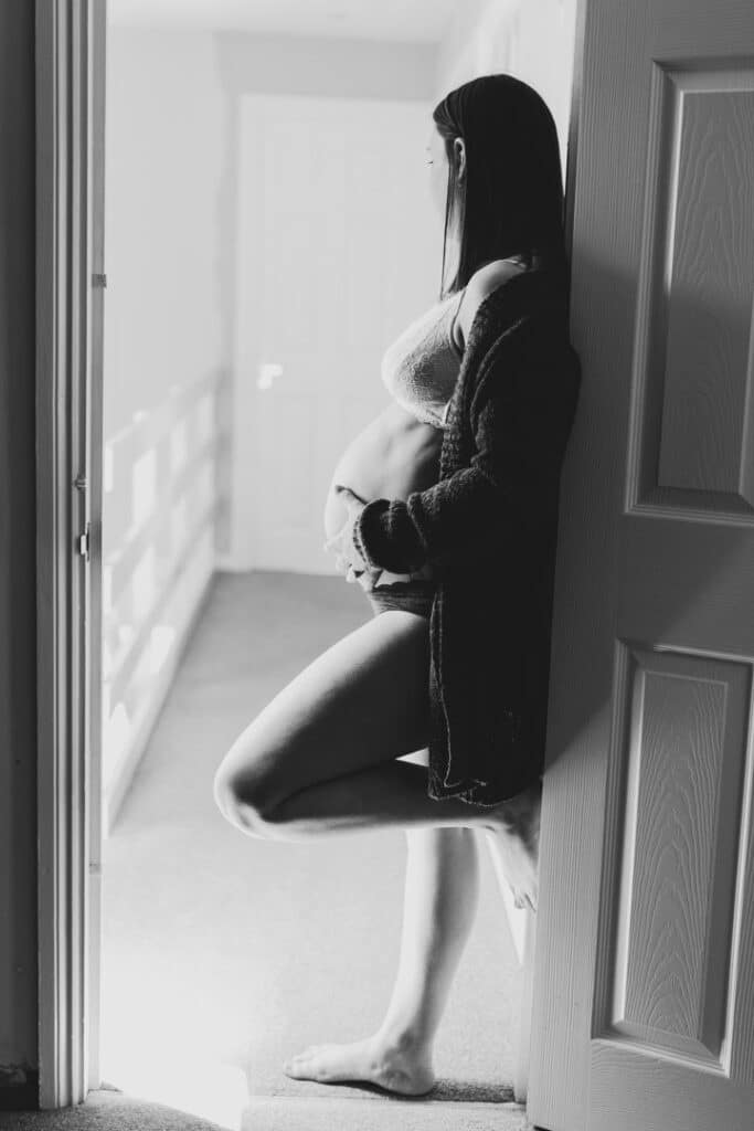 Expecting mum is standing in the door frame and she is holding her baby bump. Lovely intimate black and white maternity image. Maternity photographer in Basingstoke. Ewa Jones Photography