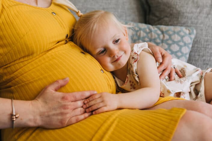 Pregnant mum is sitting on the sofa and wearing lovely yellow dress. Her older daughter is cuddling up and listening to baby bump. Maternity photographer in Hampshire. Ewa Jones Photography