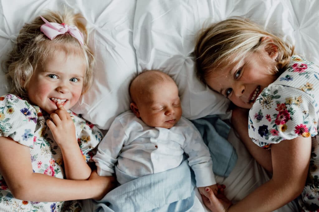 two older sisters are laying on bed and holding hands with their newborn baby brother. Newborn photographer in Hampshire. Ewa Jones Photography