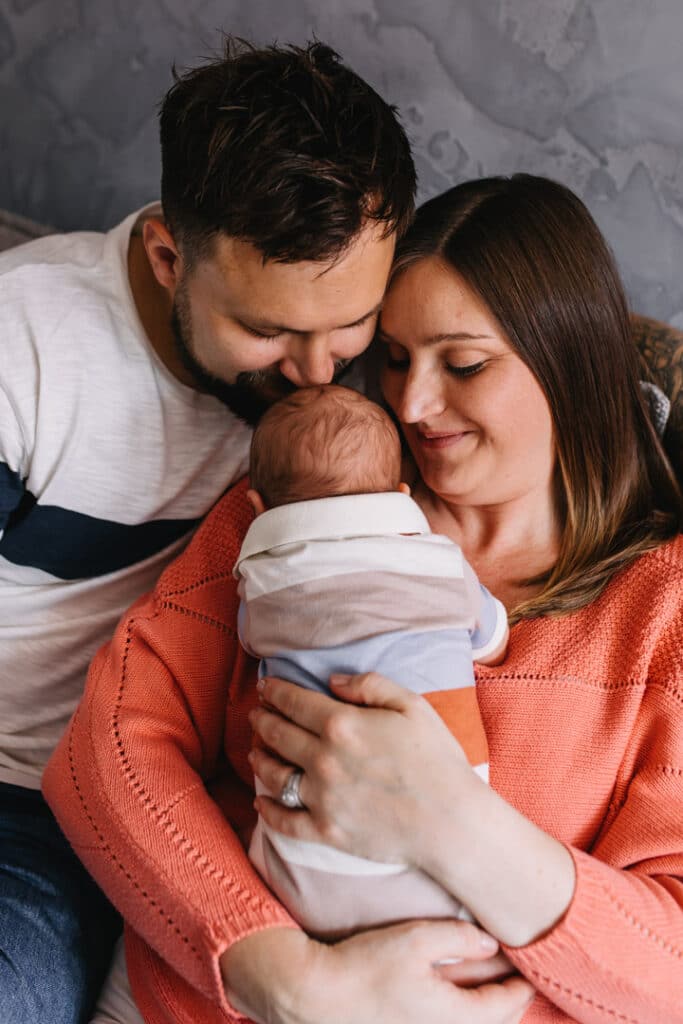 Mum and dad are sitting on the sofa and cuddling up to their newborn baby boy. Dad is kissing his newborn boy on his head. Newborn photographer in Basingstoke. Ewa Jones Photography