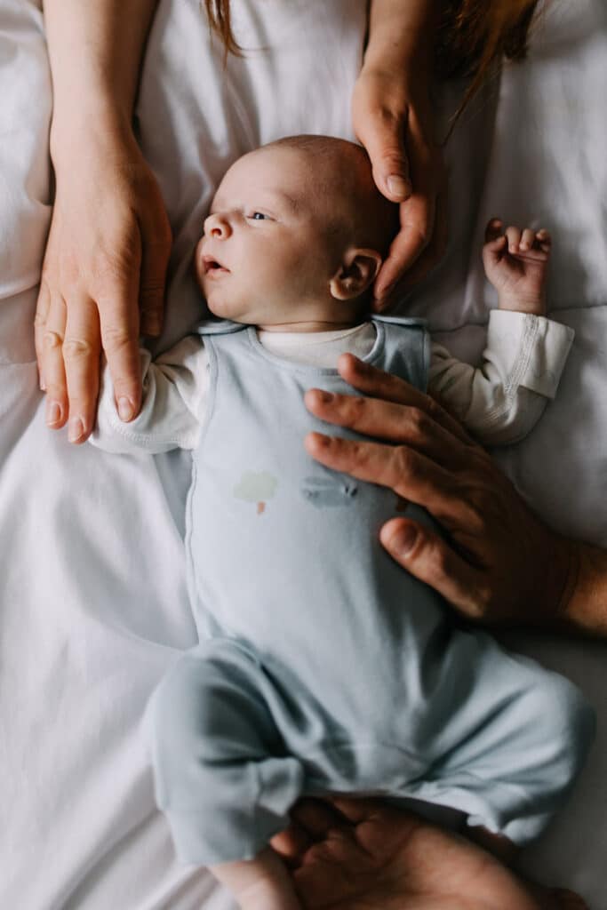 Newborn baby boy is laying on bed and looking away. His mum and dad hold his hands, head and tummy. lifestyle newborn baby photographer in Hampshire. Ewa Jones Photography