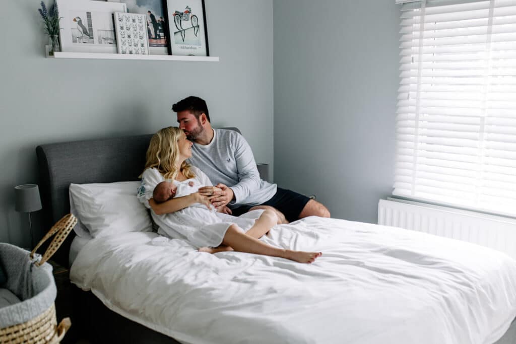 Mum and dad are sitting on the bed and dad is kissing his wife on her head. Lovely natural lifestyle newborn photo session in Hampshire. Ewa Jones Photography