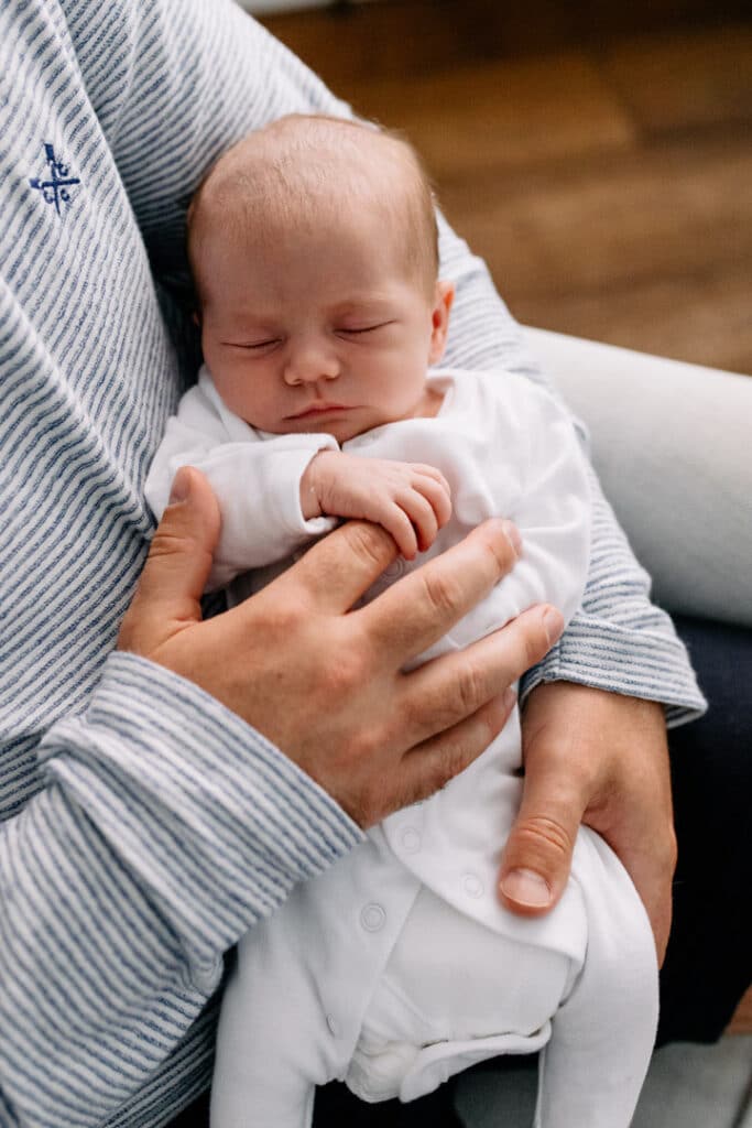 dad has his newborn baby girl in his arms and she is sleeping. Little girl is holding dads finger. Newborn photoshoot in Hampshire. Ewa Jones Photography