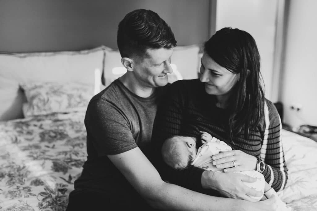 mum and dad are sitting on the bed and dad is cuddling mum and his newborn baby boy. Newborn photographer in London. Ewa Jones Photography