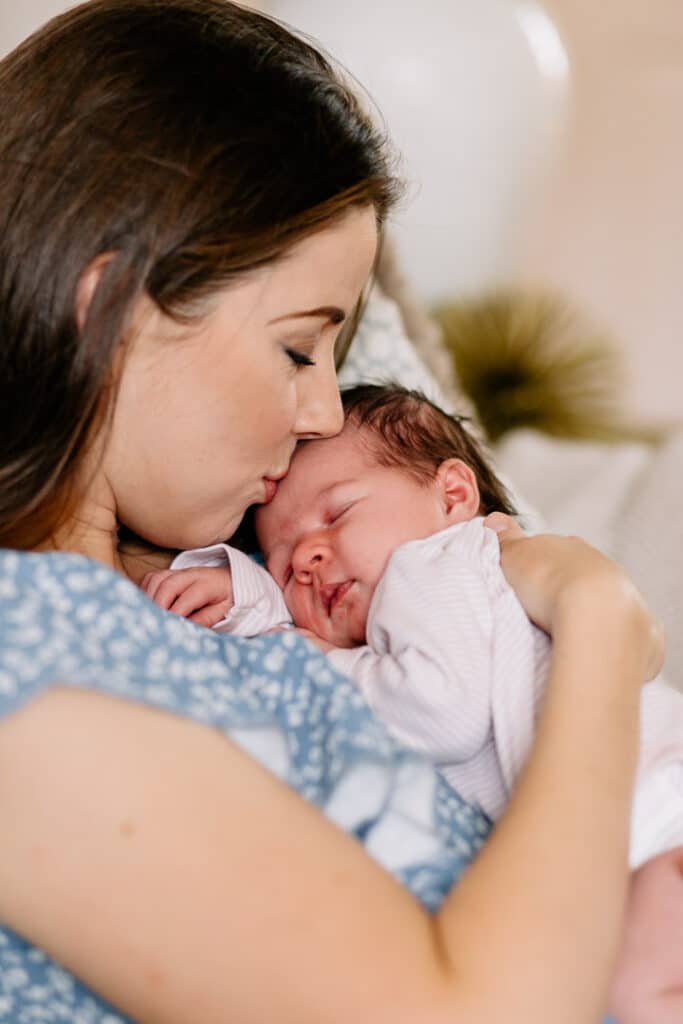 Mum is kissing her newborn baby girl on her head. Mum is sitting on the sofa and wearing lovely blue dress. Newborn photographer in Hampshire. My favourite newborn photographs of 2021. Ewa Jones Photography