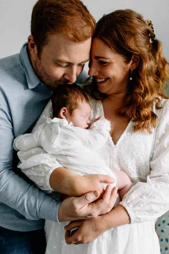 Mum and dad are cuddling baby girl. Baby girl is wearing white baby grow and dad is wearing a blue shirt. Mum is wearing white dress. Newborn photo shoot in Basingstoke, Hampshire. Ewa Jones Photography