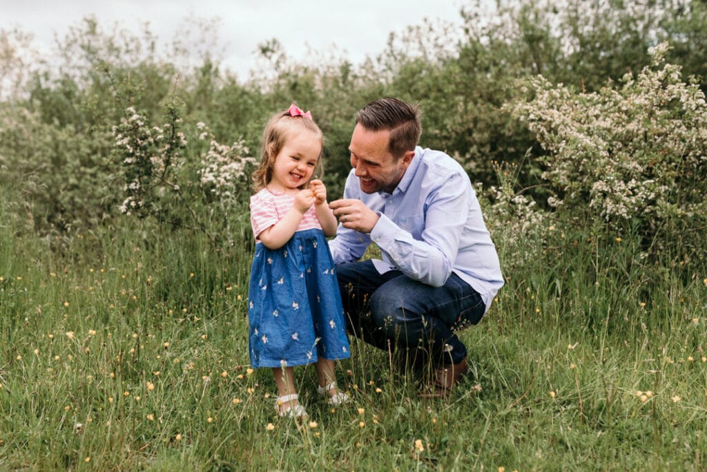 Dad is squatting and looking at his daughter. the little girl is looking at the flower and laughing. Lovely family moment of daughter and her dad. Basingstoke photographer. Ewa Jones Photography