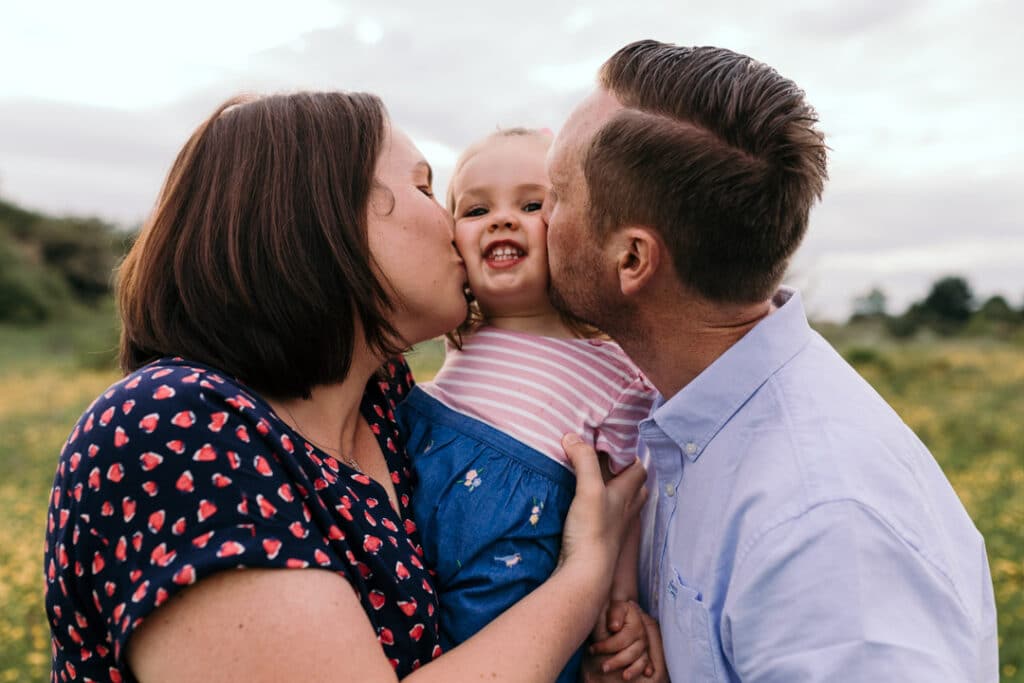 Mum and dad are kissing their daughter on her chicks. They are all wearing lovely summer outfits. family photographer in Hampshire. Ewa Jones Photography