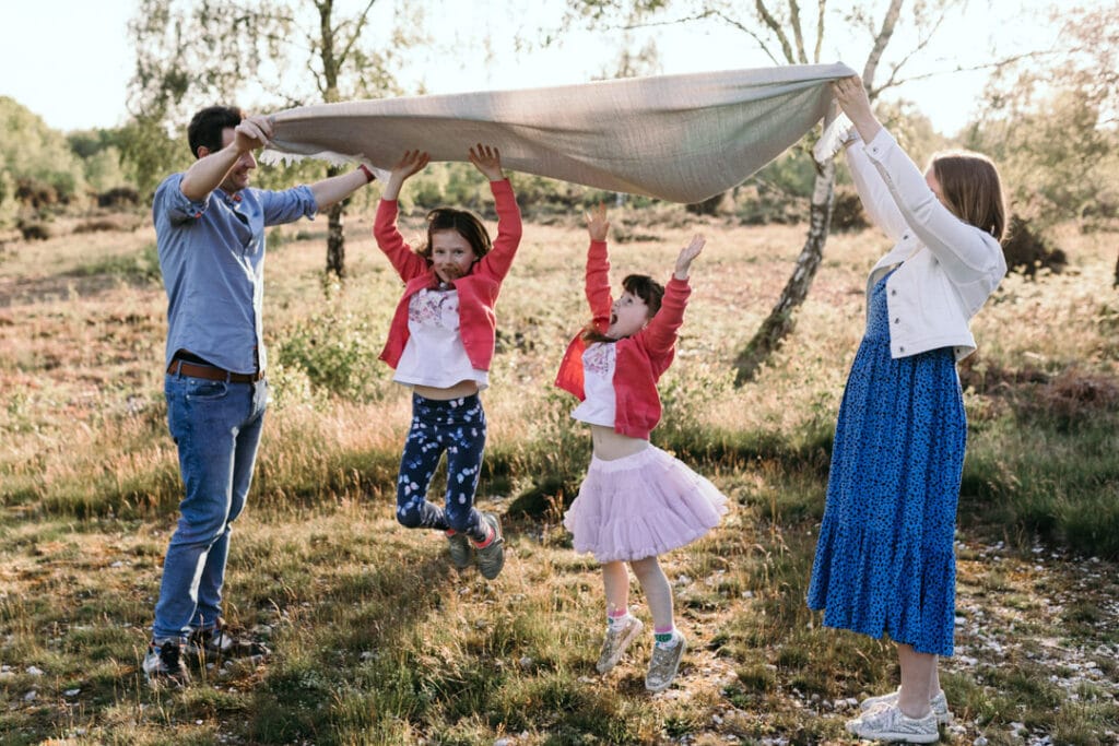 Mum and dad are holding a blanket up in the air and their daughters are jumping underneath the blanket. Family posing ideas during a family photo session. Basingstoke photographer. Ewa Jones Photography