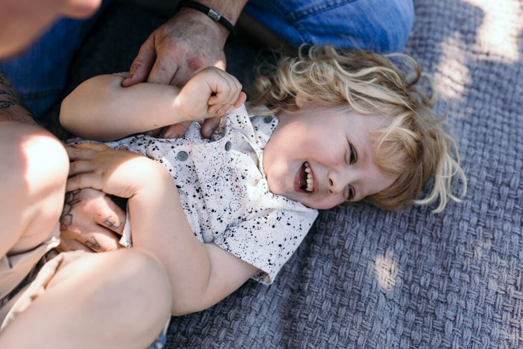 Dad is tickling his boy who is laying on the blanket. Little boy is laughing. Natural lifestyle family photographer in Hampshire. Ewa Jones Photography