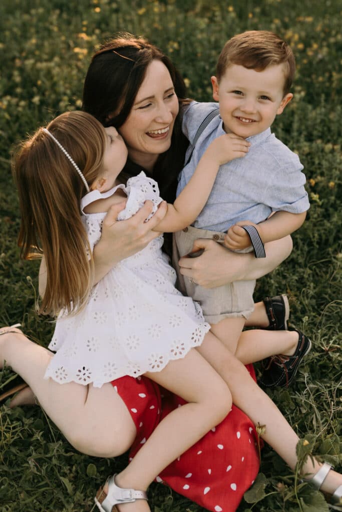 Mum is holding both her kids and smiling. Little boy is looking at the camera and little girl is kissing her mum. Family photographer in Basingstoke, Hampshire. Ewa Jones Photography