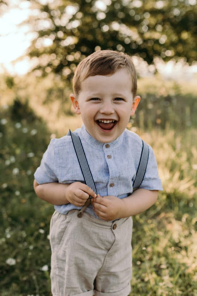 Little boy is looking at the camera and laughing. His is wearing lovely blue t-shirt and grey linen shorts. Family photo shoot in summer in Basingstoke. Ewa Jones Photography