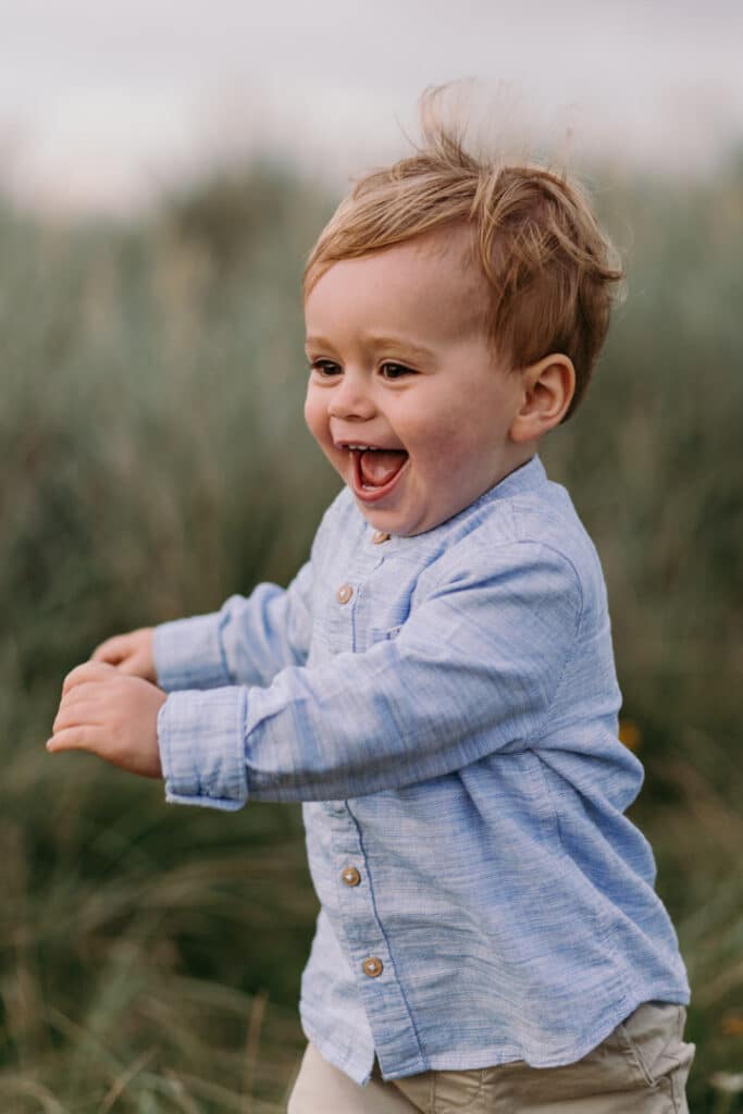 Little boy is running and laughing. He is wearing lovely blue shirt. Summer family session at the beach in Hampshire. Ewa Jones Photography
