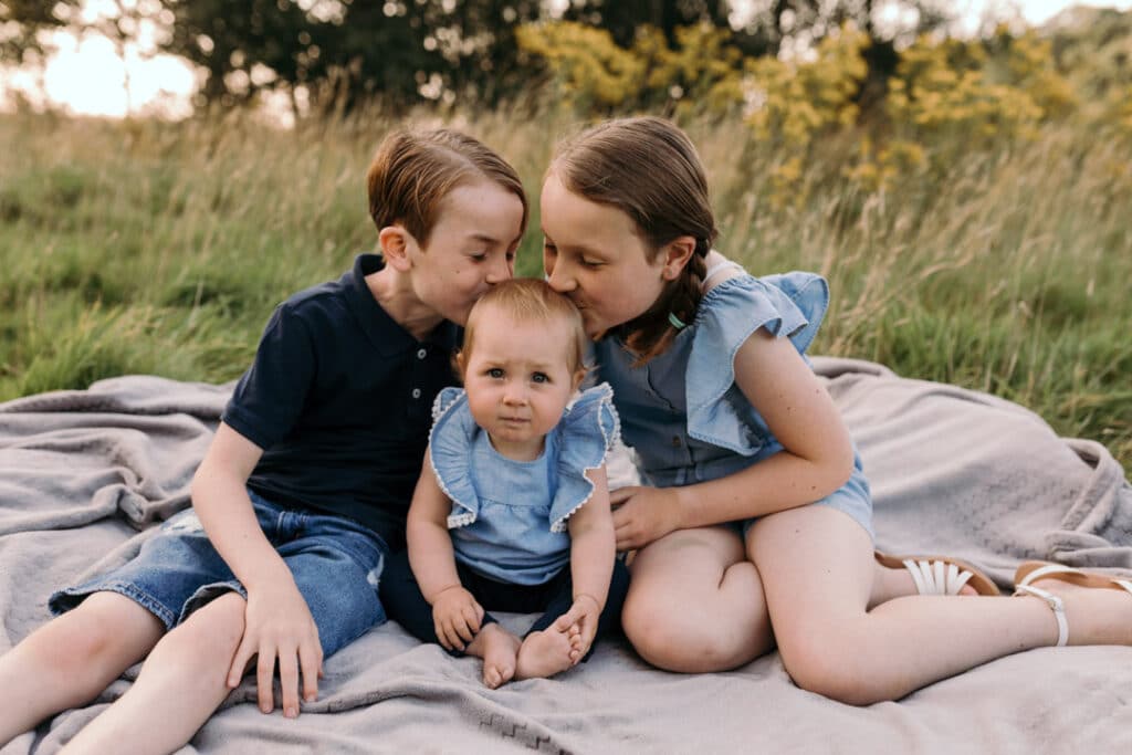 Three siblings are sitting on the blanket at older brother and sister are kissing their baby girl. Summer family photo session in Basingstoke, Hampshire. Ewa Jones Photography