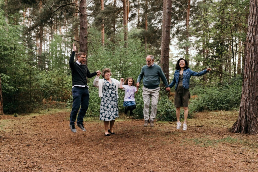 Family is holding hands and jumping in the air. Lovely family connection. They are in the woods with lovely trees behind them. Family photographer in Basingstoke, Hampshire. Ewa Jones Photography