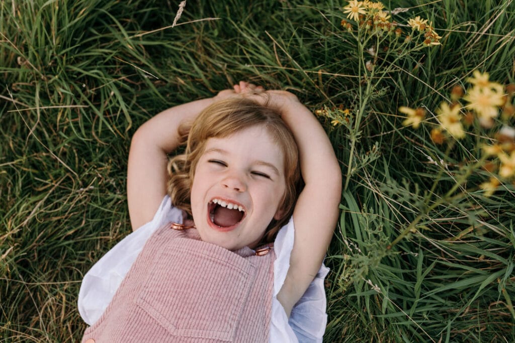 Little girl is laying on the grass with her hands up. she is laughing. Summer family session in Basingstoke, Hampshire. Ewa Jones Photography