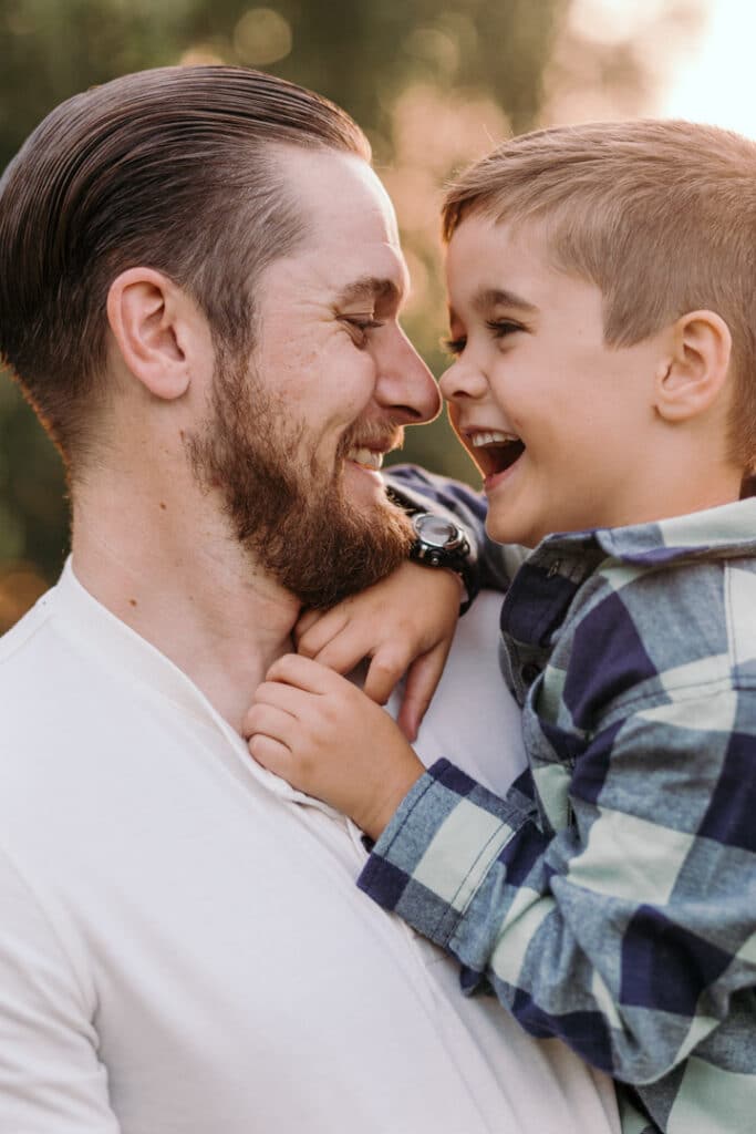 dad and son are laughing. Son is wearing a checked shirt and dad is wearing a white top. Family sunset photo session in Basingstoke, Hampshire. Ewa Jones Photography