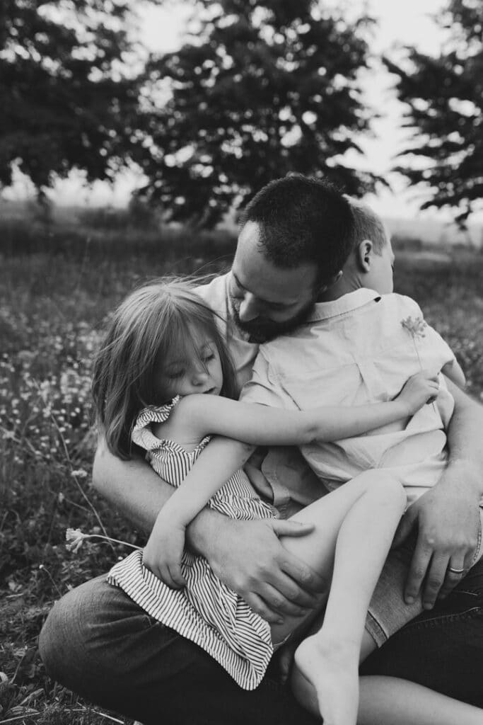 Black and white image of dad cuddling his children. They sit on the grass and dad is looking down at his daughter. Family photographer in Basingstoke. Ewa Jones Photography