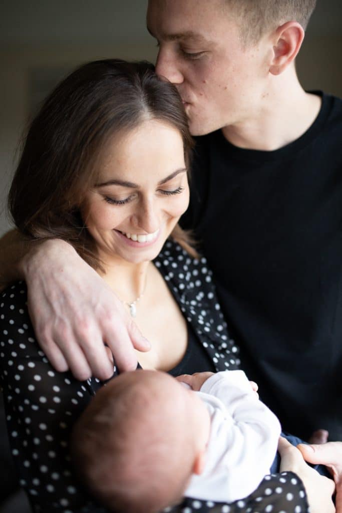 Mum and dad are standing close to each other and dad is kissing mums head. She is holding her newborn baby. Newborn photographer in Hampshire. Hampshire photographer. Ewa Jones Photography