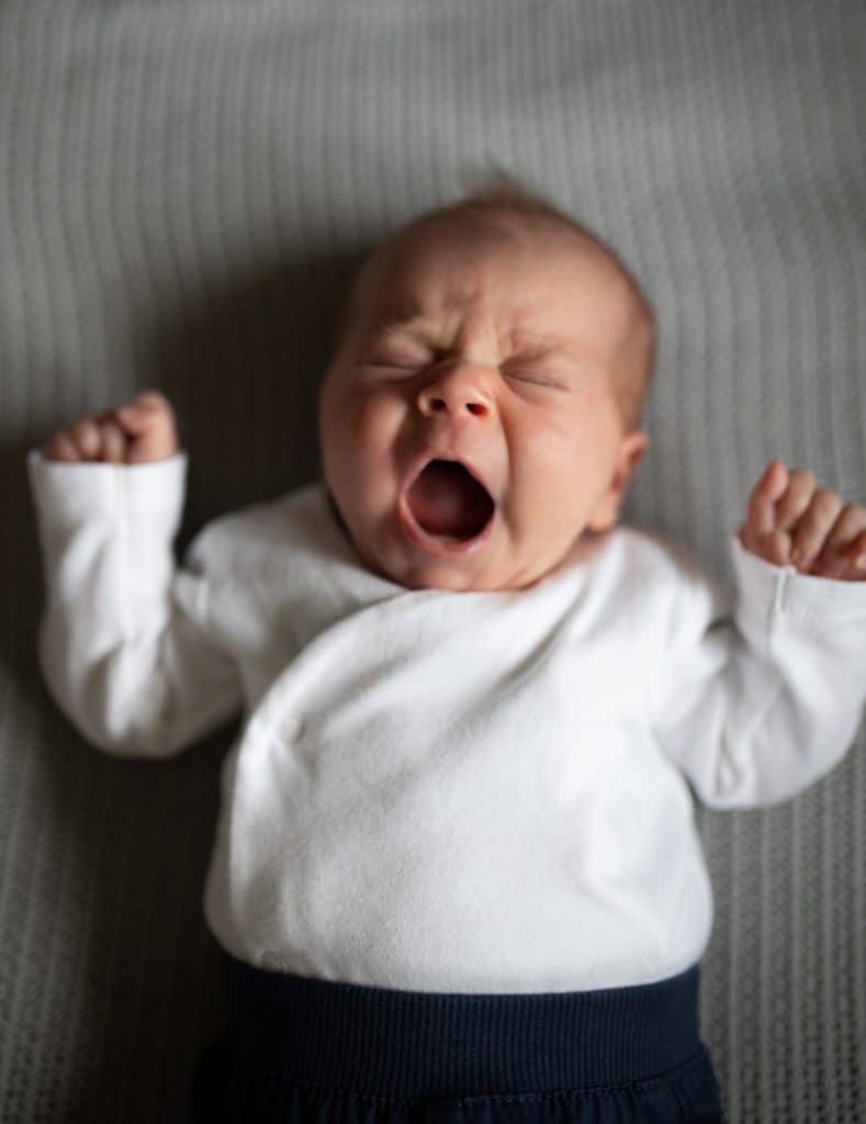 Newborn baby boy is laying on the blanket and yawning. he is wearing white body and dark trousers. Newborn photo session in Hampshire. Hampshire newborn photographer. Ewa Jones Photography