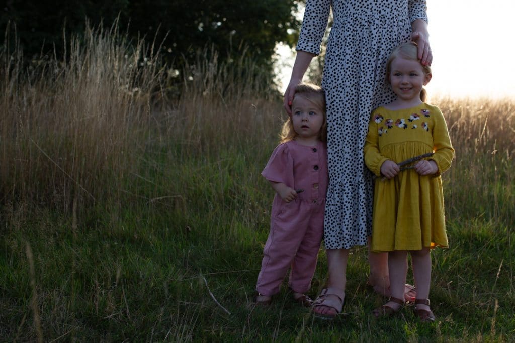 Mum and her three daughters are standing in the field of wild flowers and grass. family photo shoot in Hampshire. Basingstoke photographer. little baby boy is looking at the camera and smiling. Family photographer in Basingstoke, Hampshire. Ewa Jones Photography