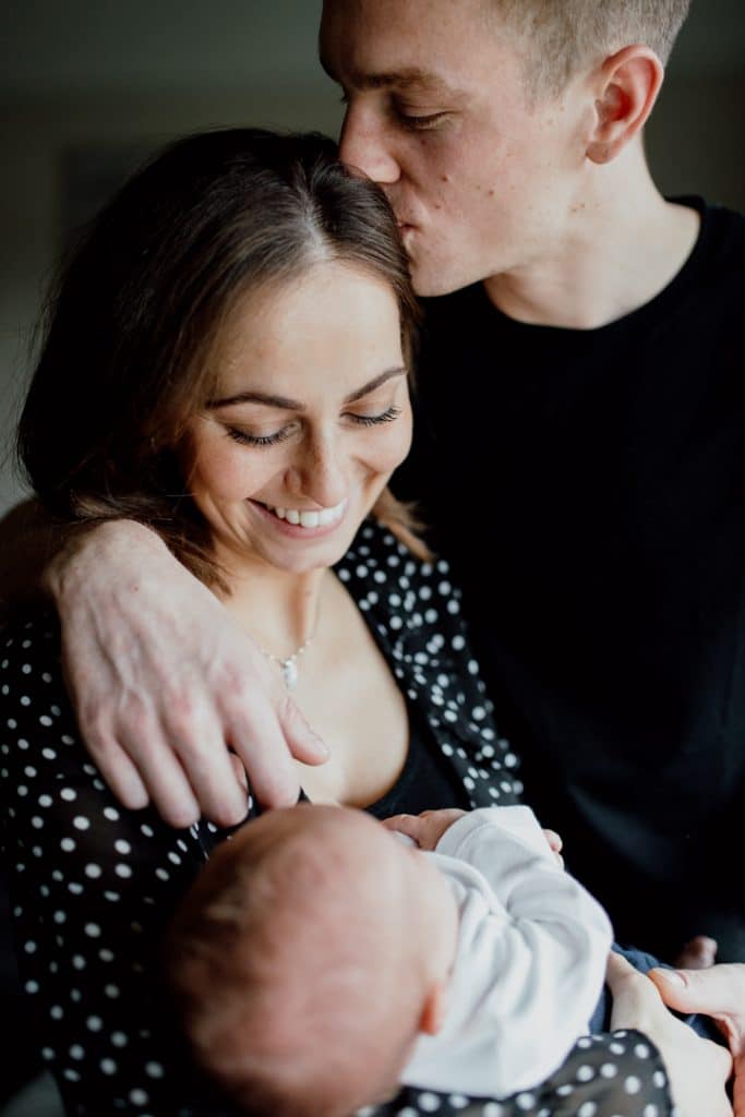 Mum and dad are standing close to each other and dad is kissing mums head. She is holding her newborn baby. Newborn photographer in Hampshire. Hampshire photographer. Ewa Jones Photography