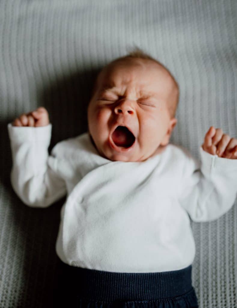 Newborn baby boy is laying on the blanket and yawning. he is wearing white body and dark trousers. Newborn photo session in Hampshire. Hampshire newborn photographer. Ewa Jones Photography