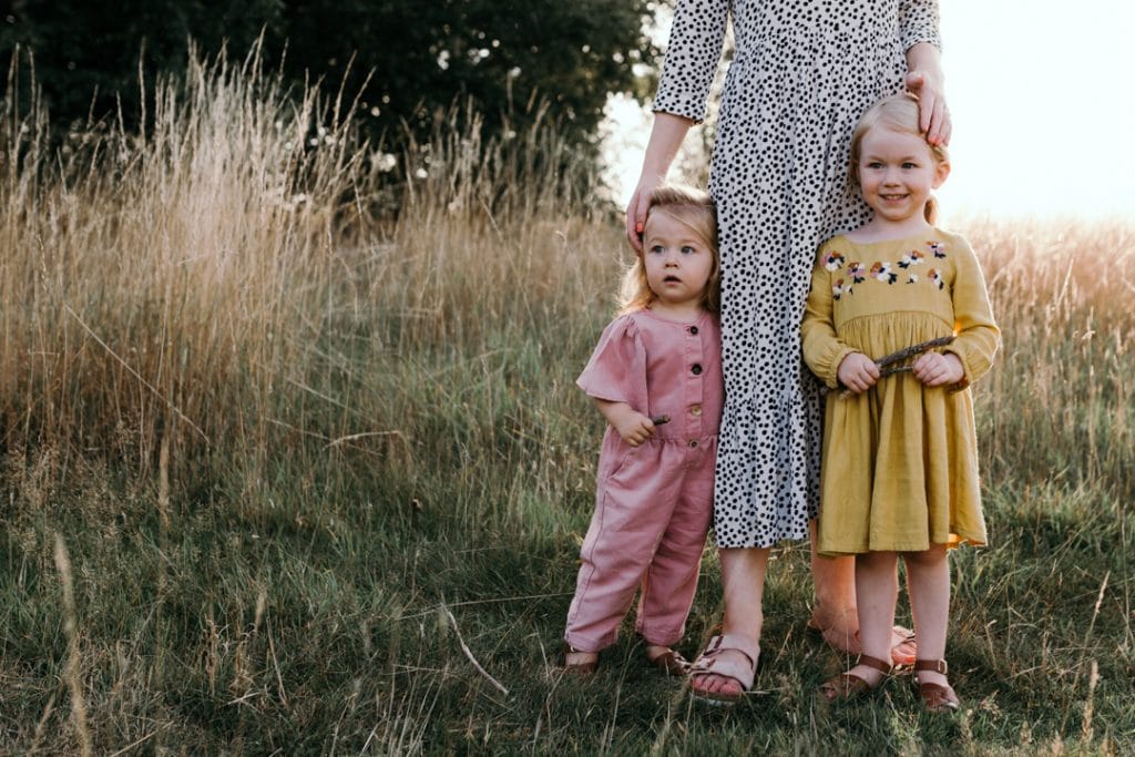 Mum and her three daughters are standing in the field of wild flowers and grass. family photo shoot in Hampshire. Basingstoke photographer. little baby boy is looking at the camera and smiling. Family photographer in Basingstoke, Hampshire. Ewa Jones Photography