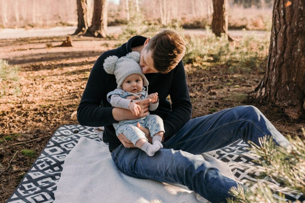 Dad is kissing his baby boy on his head and sitting on the blanket. Family photographer in Basingstoke, Hampshire. Ewa Jones Photography