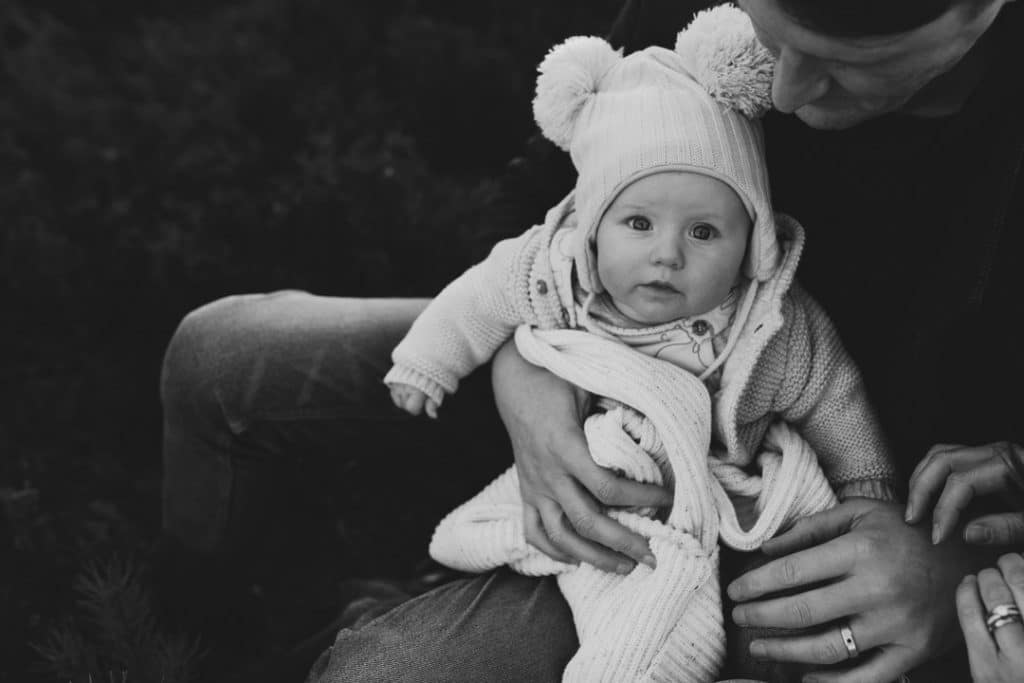 Black and white image of a baby boy sitting on daddy's lap and looking at the camera. Family photographer in Hampshire. Ewa Jones Photography