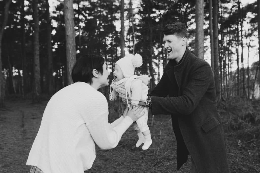 Mum and dad are laughing and their little boy is lifted in the air by dad. Fun family photo shoot.Outdoor family photographer in Hampshire. Ewa Jones Photography